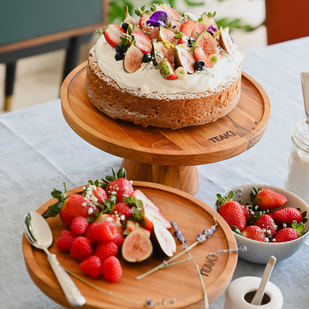 Round teakwood cake stand showcasing a berry-topped cream cake on a light tablecloth with fresh fruit.