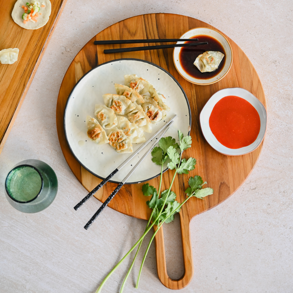 Teakwood round serving board with a handle, topped with a plate of dumplings, sauces, and fresh cilantro.