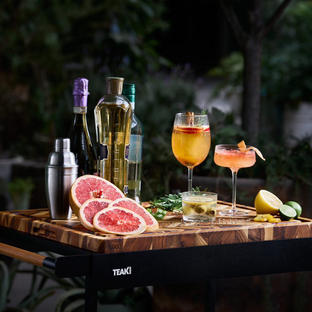 Extra-large teakwood herringbone cutting board styled with cocktails and sliced grapefruit on a black bar cart.