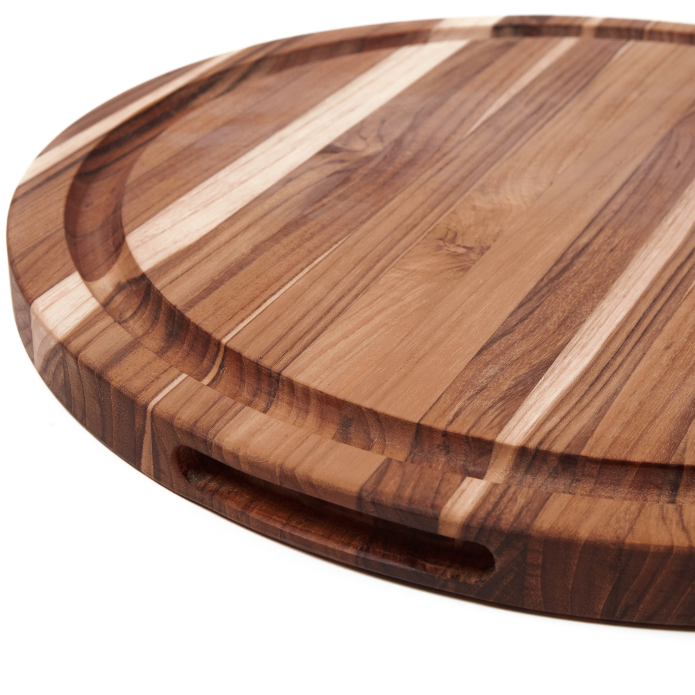 Round teakwood carving board showing its deep juice groove, side handle, and rich edge grain patterns.