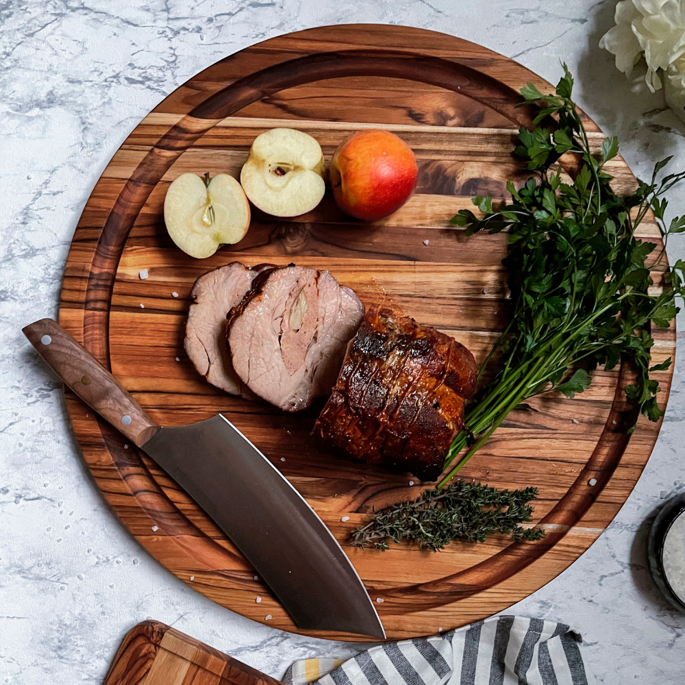 Round teakwood carving board with juice groove, styled with sliced roasted meat, herbs, and apples.
