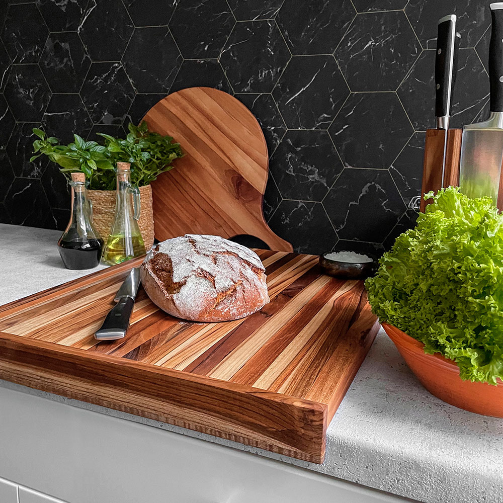 Large edge grain teakwood cutting board with an integrated lip, featuring a rustic bread loaf in a modern kitchen.