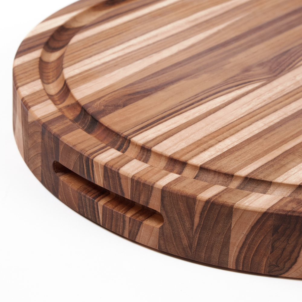 Close-up showing the edge grain and juice groove of a round teakwood carving board with a side handle.
