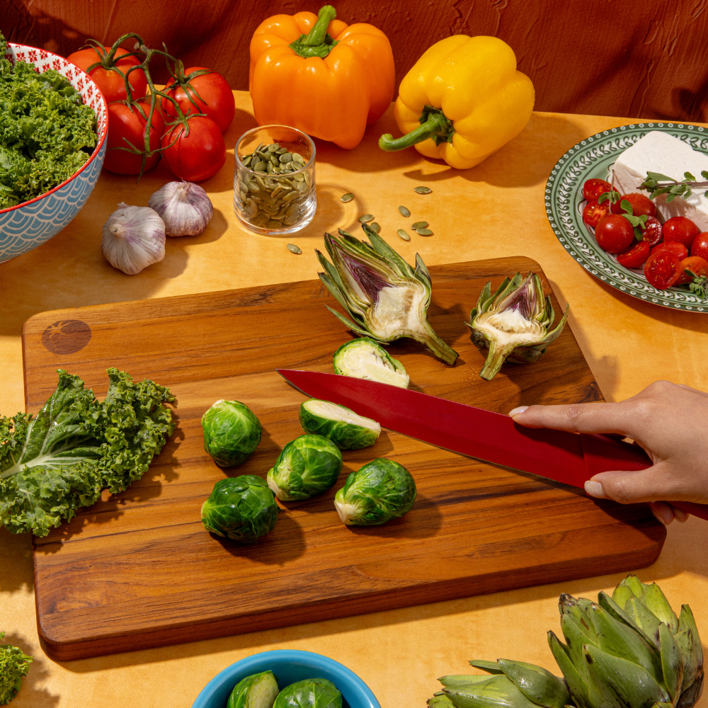 Rectangular teakwood cutting board with rich wood grain used for slicing brussels sprouts with a red knife.