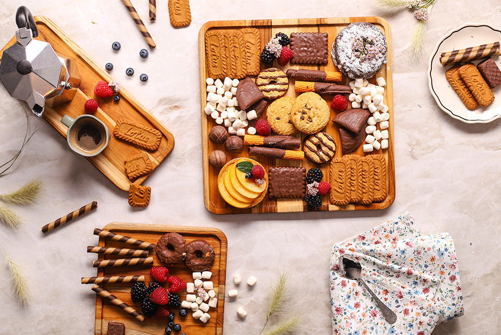 Three sustainable teakwood serving trays displaying an assortment of sweets and berries on a marble tabletop.