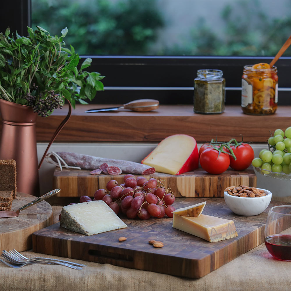 Long end grain teakwood cheese board topped with various cheeses and grapes in a rustic kitchen setting.