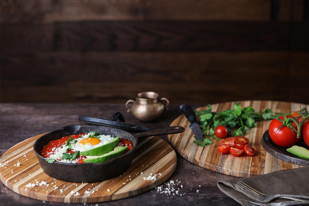 Round edge grain teakwood cutting board used as a serving platter for a cast iron skillet and fresh ingredients.
