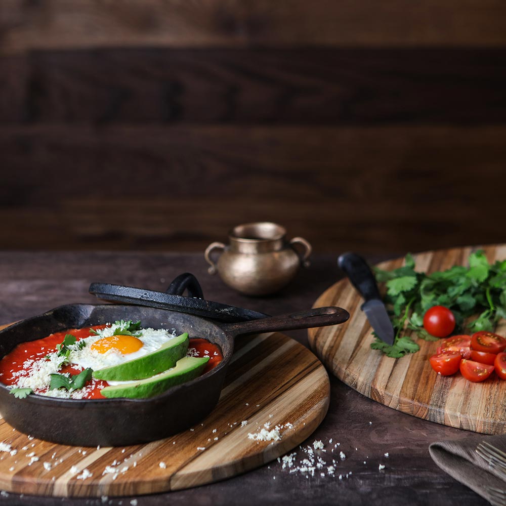 Round edge grain teakwood cutting board used for serving a cast iron skillet meal and prepping fresh vegetables.