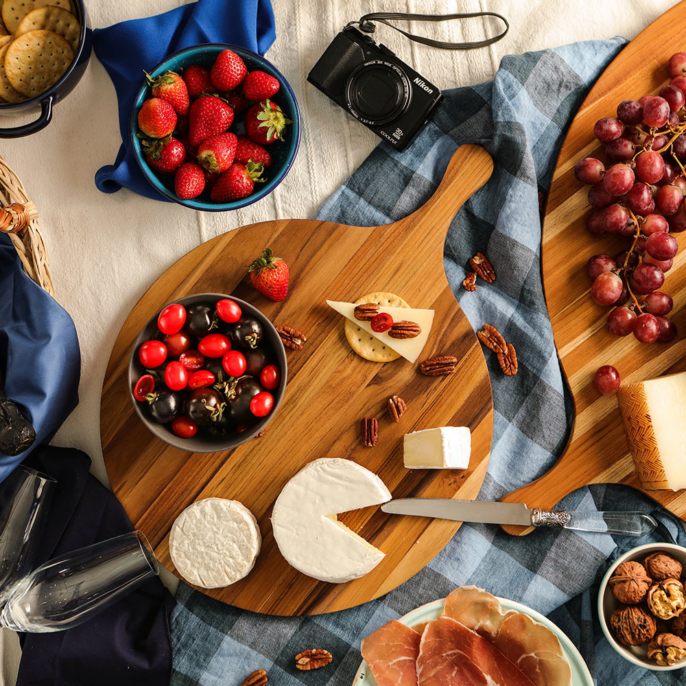 Round edge grain Atlas teakwood pizza serving board with handle, styled with brie cheese and crackers for charcuterie.