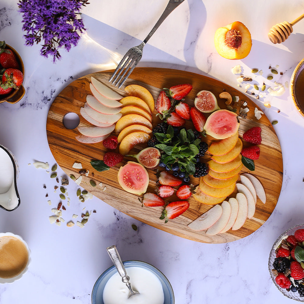 Elegant oval teak serving board topped with fresh sliced fruits on a white marble surface.