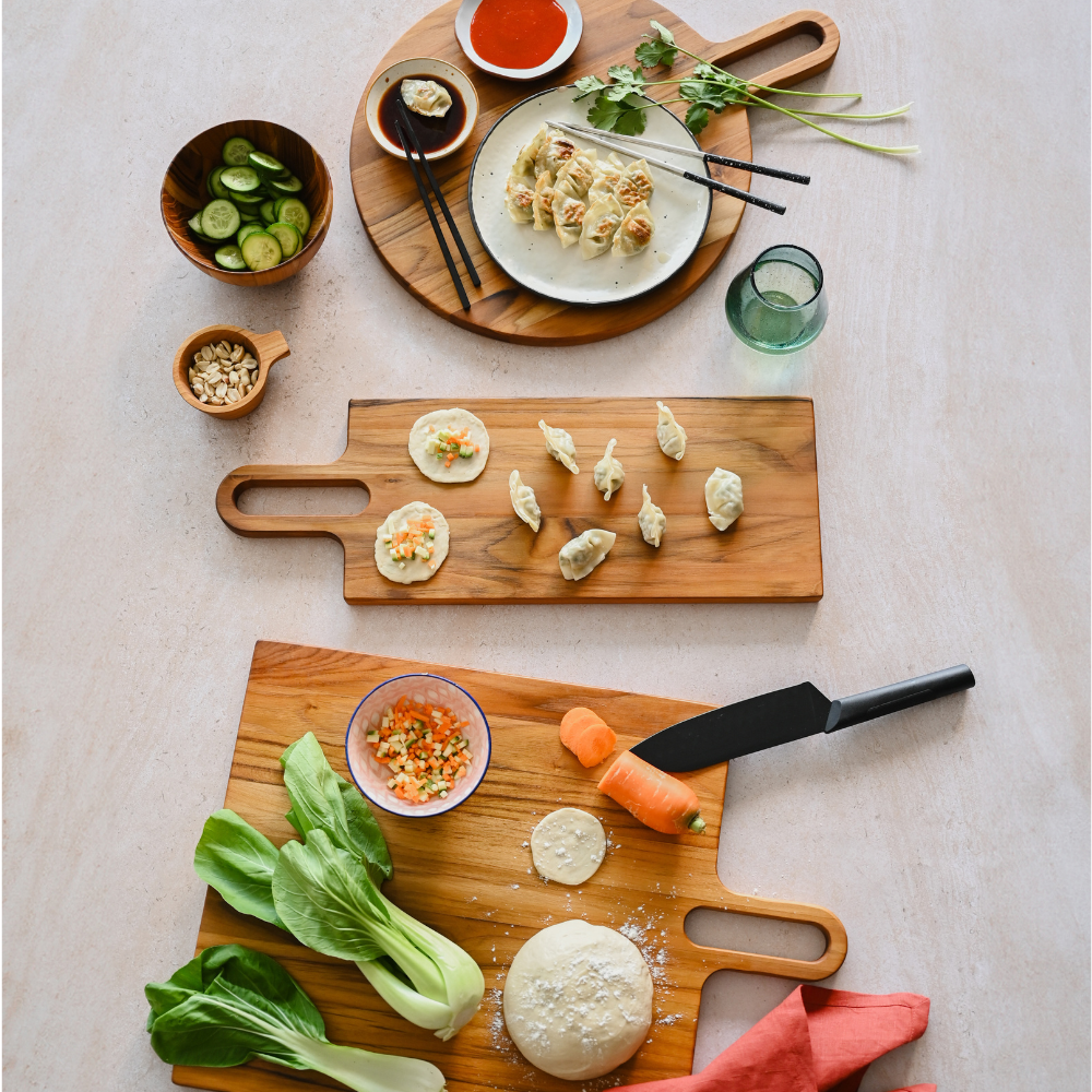 Long rectangular teakwood serving board with a handle, holding dumplings in a lifestyle kitchen scene.