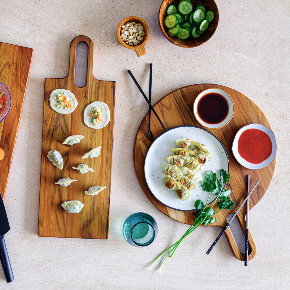 Round teakwood serving board holding a plate of dumplings and dipping sauces on a light-colored surface.