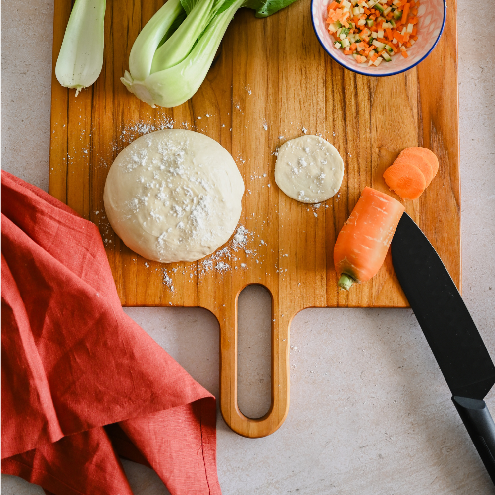 Large square teakwood serving board with a handle, used for food prep with dough and fresh vegetables.