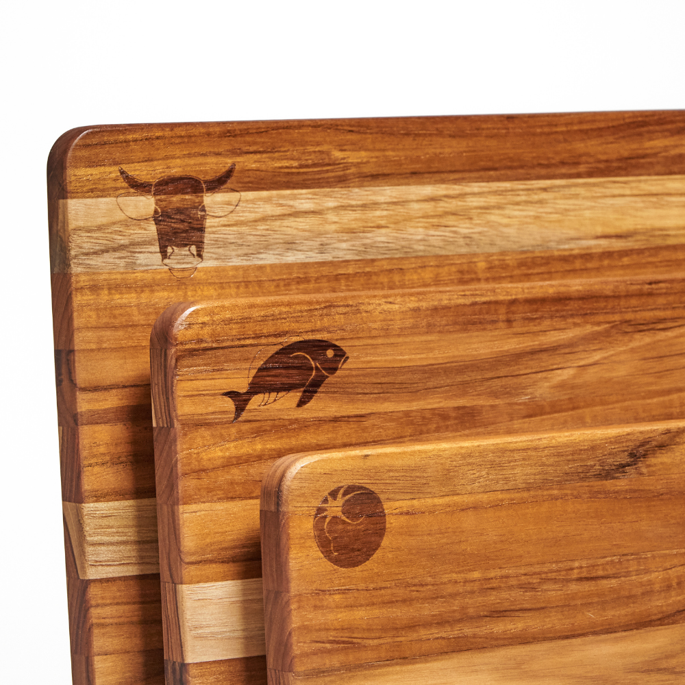 Stacked teakwood kitchen boards with decorative cow, fish, and tomato engravings showcasing natural wood grain.