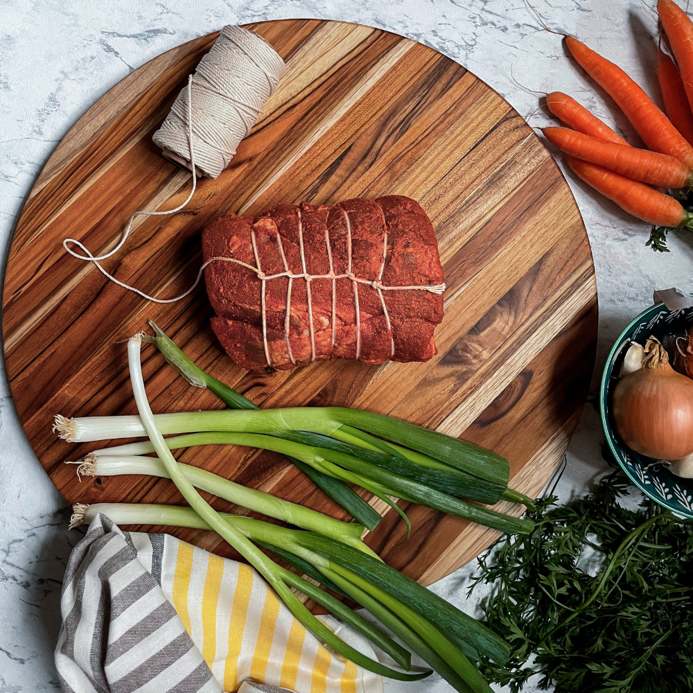 Round edge grain teakwood carving board featuring a tied meat roast and green onions in a kitchen setting.