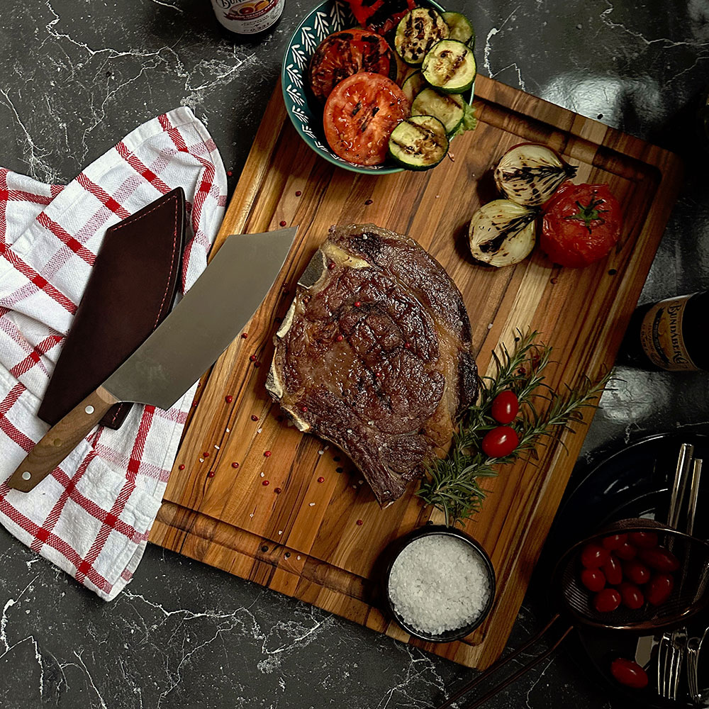Large edge grain teakwood BBQ board featuring a grilled steak and roasted vegetables on a dark marble surface.