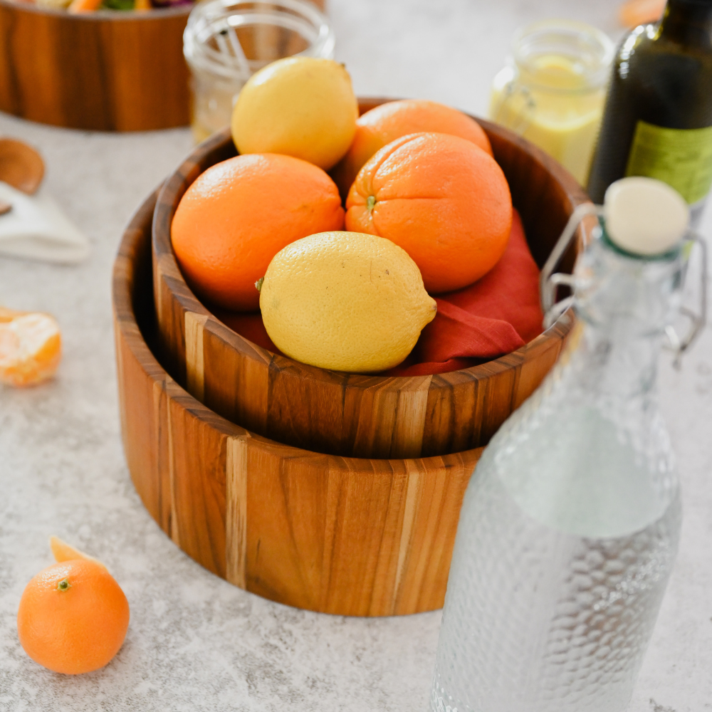 Stacked teakwood salad bowls with natural grain, filled with fresh lemons and oranges in a kitchen setting.