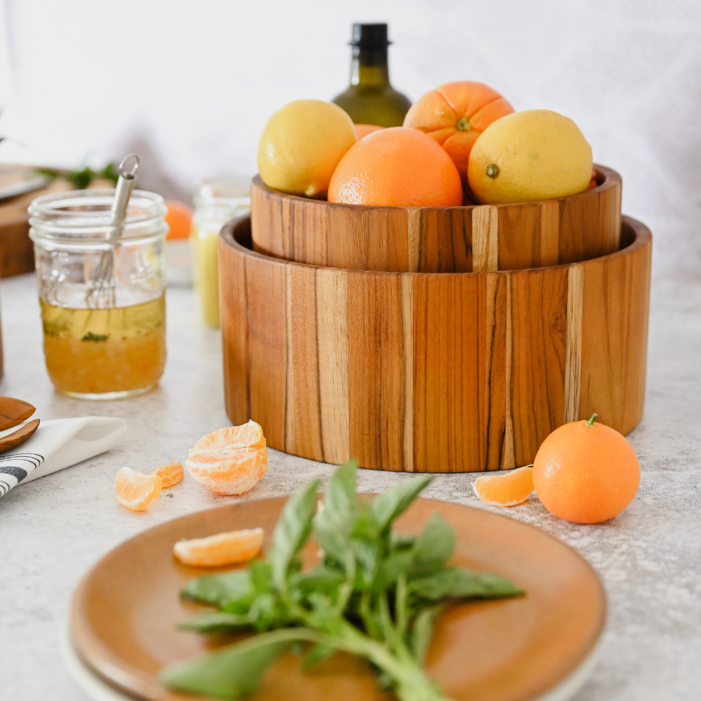 Nested teakwood salad bowls filled with fresh citrus fruit on a kitchen counter with herbs and dressing.