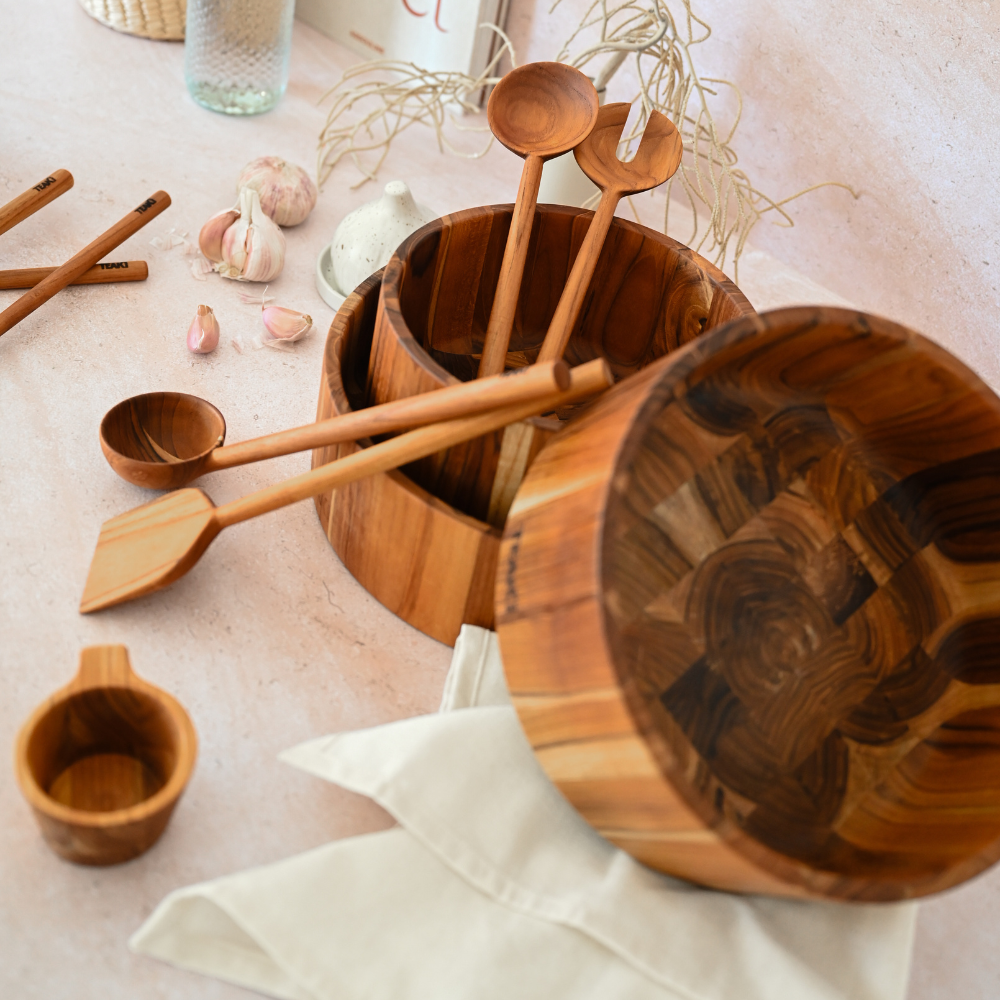 Sculptural teakwood salad bowl and matching wooden serving utensils on a light surface with fresh garlic.