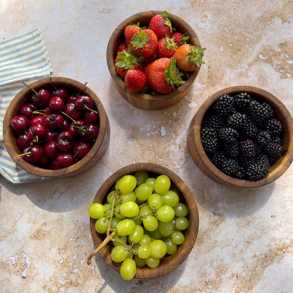 Set of four round teakwood bowls filled with fresh berries and grapes on a neutral stone surface.