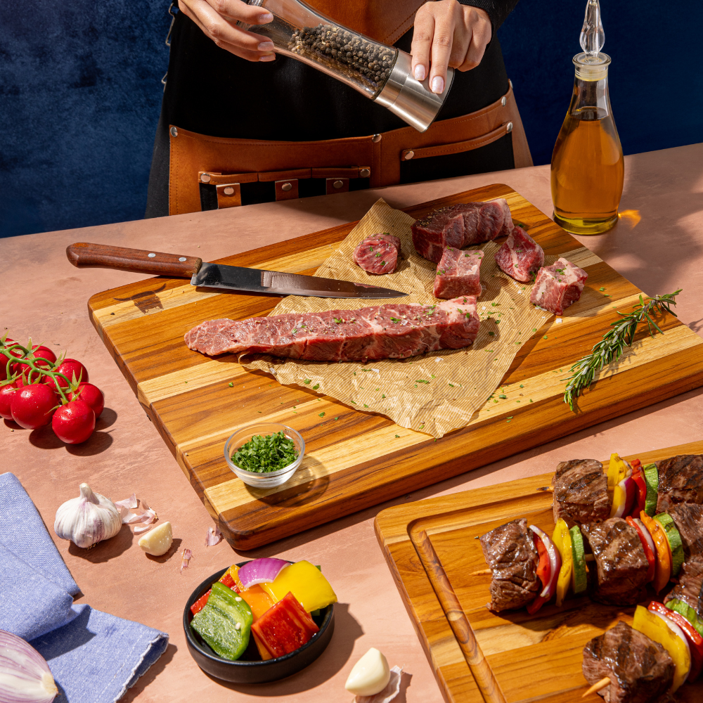 Rectangular teakwood kitchen board used for prepping raw steak with fresh vegetables and a pepper grinder.