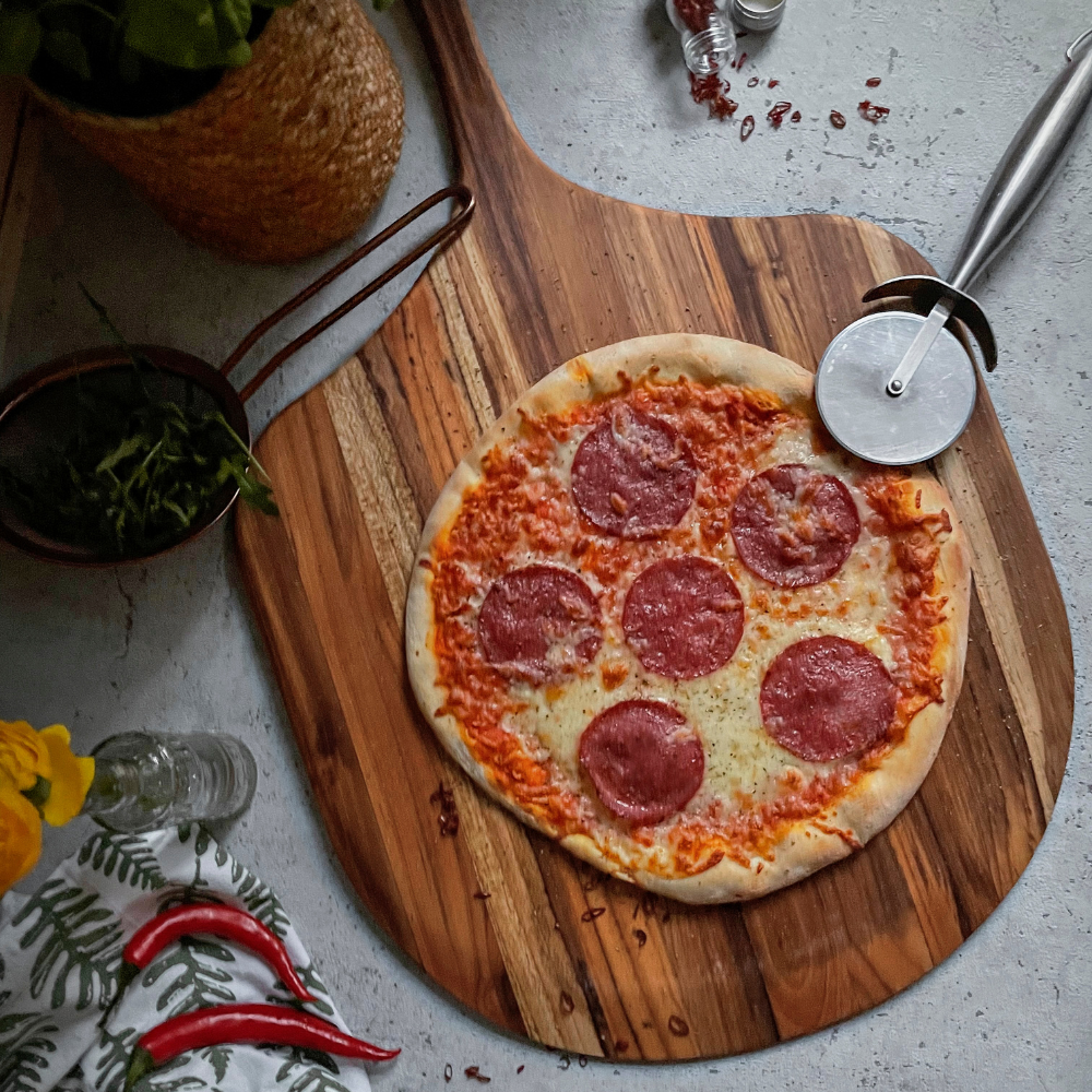 Canoe paddle teakwood pizza peel used as a serving board for a pepperoni pizza with a stainless steel cutter.