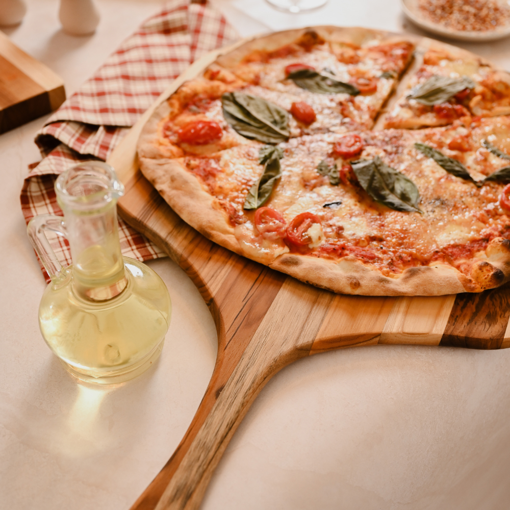 Small teakwood canoe paddle pizza peel holding a fresh tomato and basil pizza beside an oil carafe.