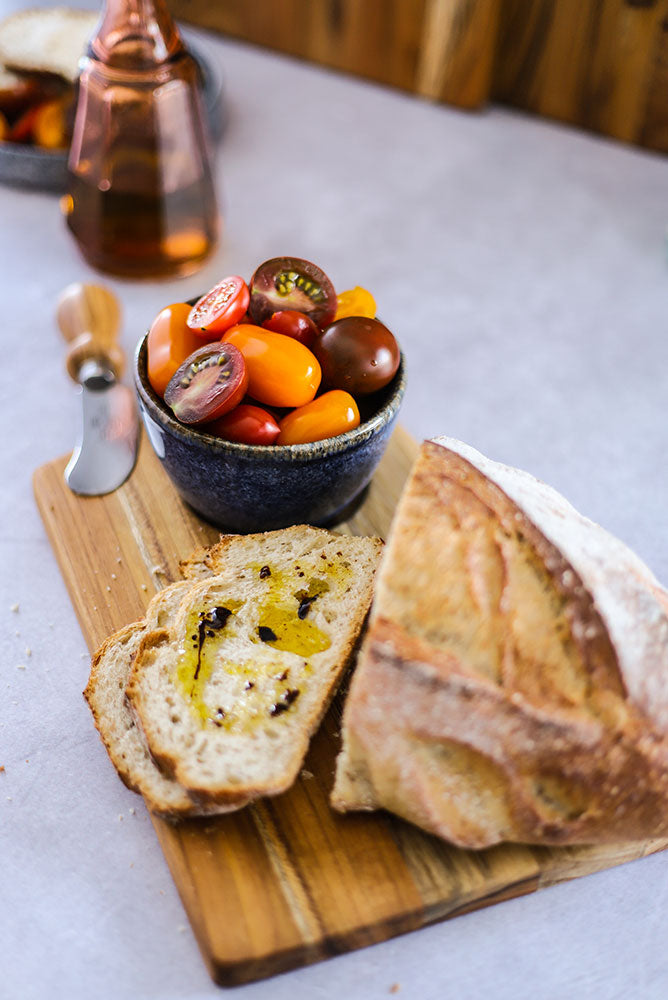 Teakwood trapeze serving board holding crusty bread with olive oil and a bowl of fresh tomatoes.