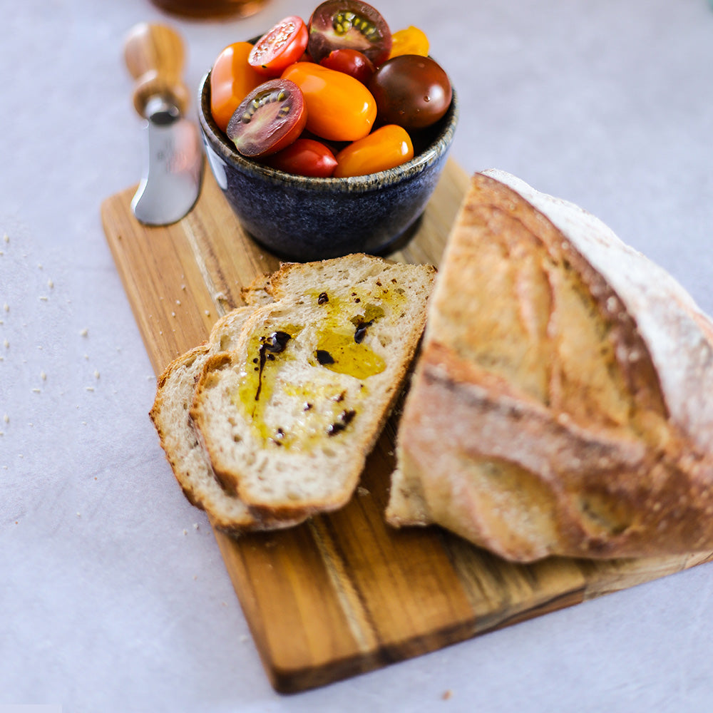 Small trapeze teak serving board holding crusty bread with olive oil and a bowl of fresh cherry tomatoes.
