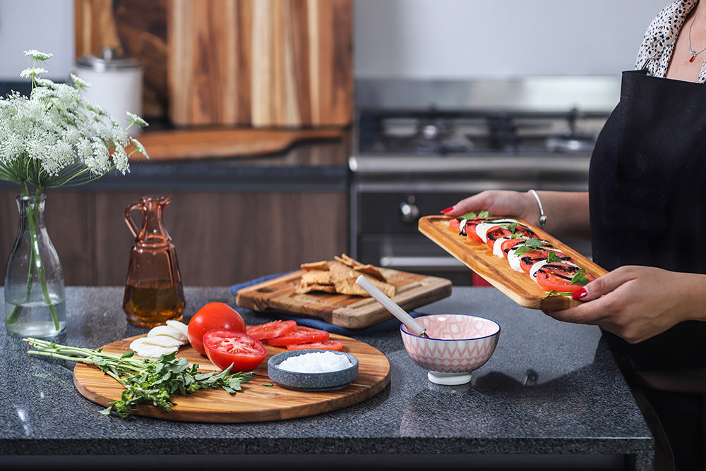 Rectangular teak serving tray featuring a Caprese salad, held by a person in a modern kitchen setting.