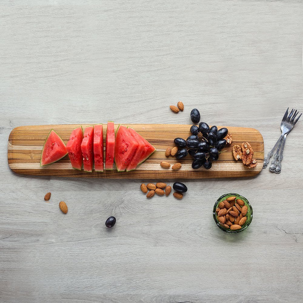Long teakwood serving board with rounded edges, featuring sliced watermelon and grapes on a light grey wood surface.