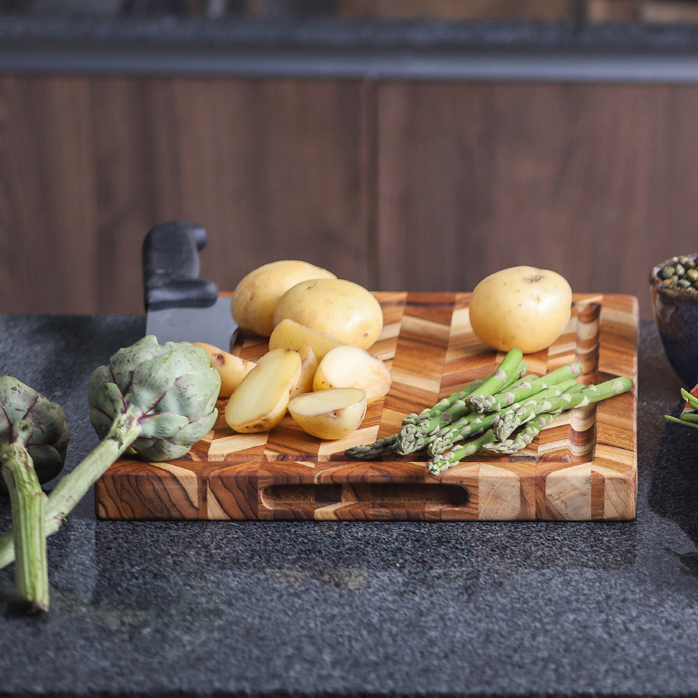 Herringbone teakwood cutting board with potatoes and asparagus on a dark granite kitchen countertop.
