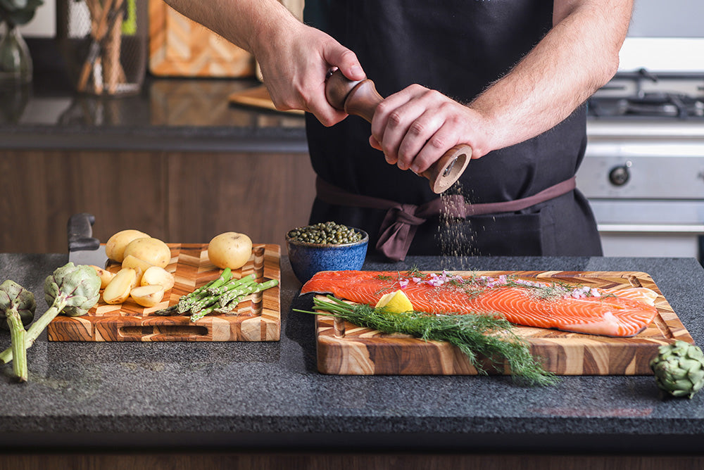 Teakwood herringbone cutting board used for seasoning salmon and preparing vegetables in a kitchen.