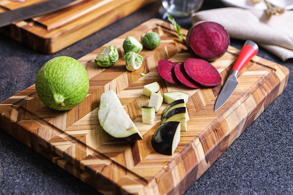 Sustainable teakwood herringbone cutting board featuring sliced vegetables and a red paring knife.