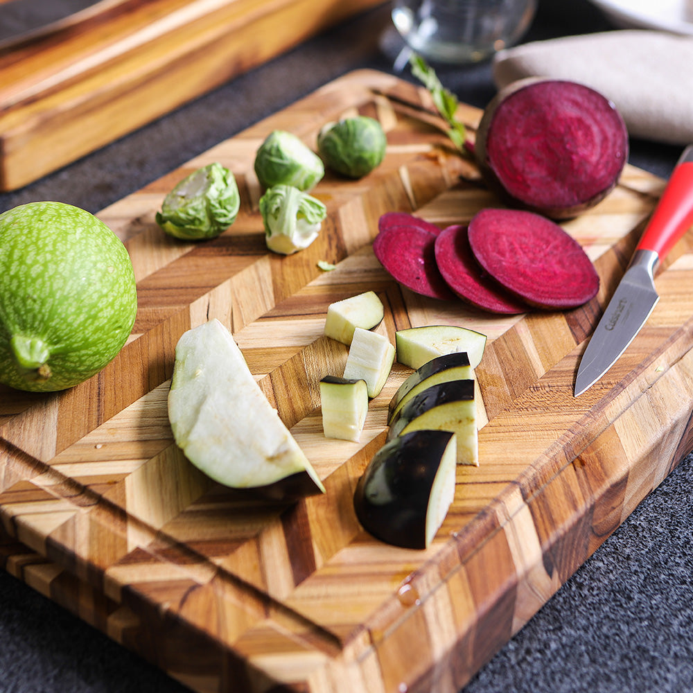 Herringbone teakwood cutting board topped with sliced beets, eggplant, and fresh vegetables.