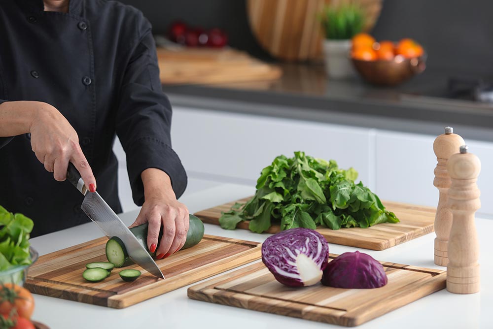 Chef slicing vegetables on rectangular teak wood cutting boards with juice canals in a bright kitchen.