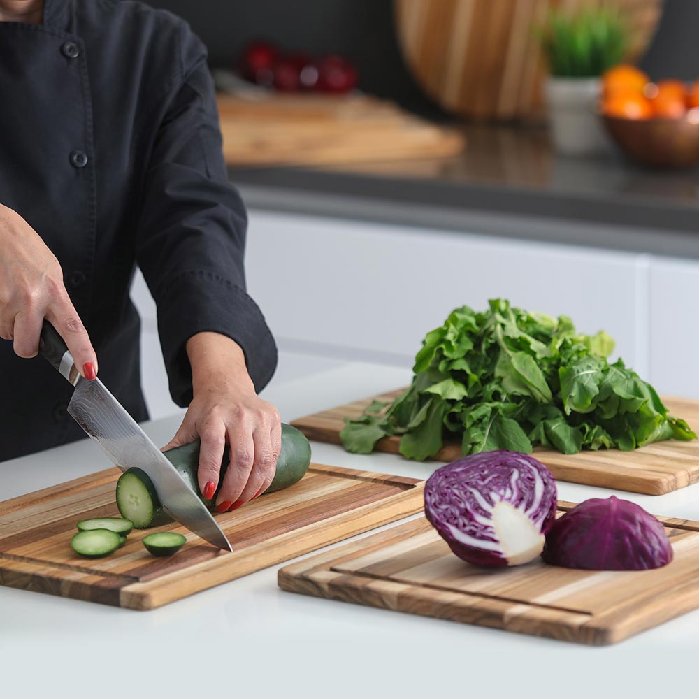 Teakwood edge grain cutting boards with juice canals used to slice fresh cucumber and cabbage in a kitchen.