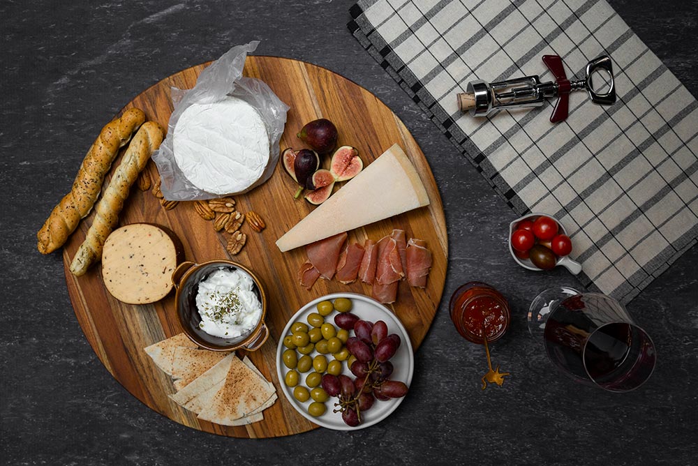 Round teak wood board styled with a charcuterie spread of cheeses and fruits on a dark surface.