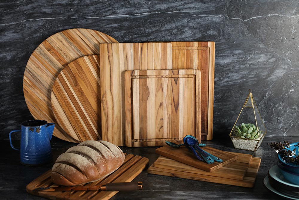Teakhaus Essential Collection round and rectangular teakwood cutting boards displayed on a dark marble surface.
