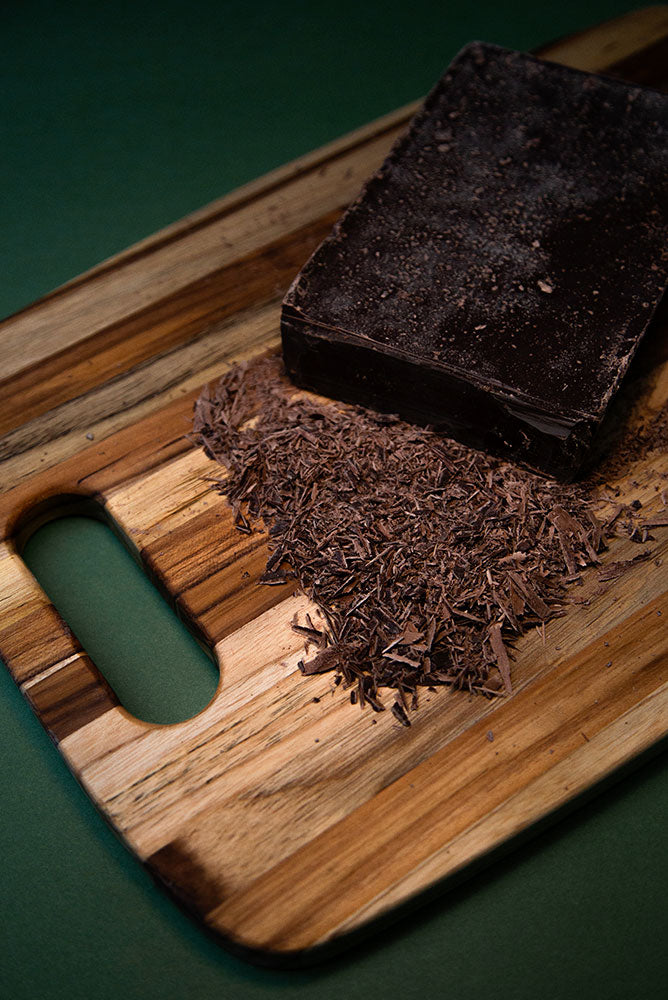Teak wood edge grain chopping board with a hole handle, featuring a block of dark chocolate and fine shavings.