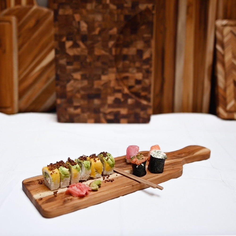 Slim teak paddle serving board featuring an assortment of sushi and chopsticks on a white tablecloth.