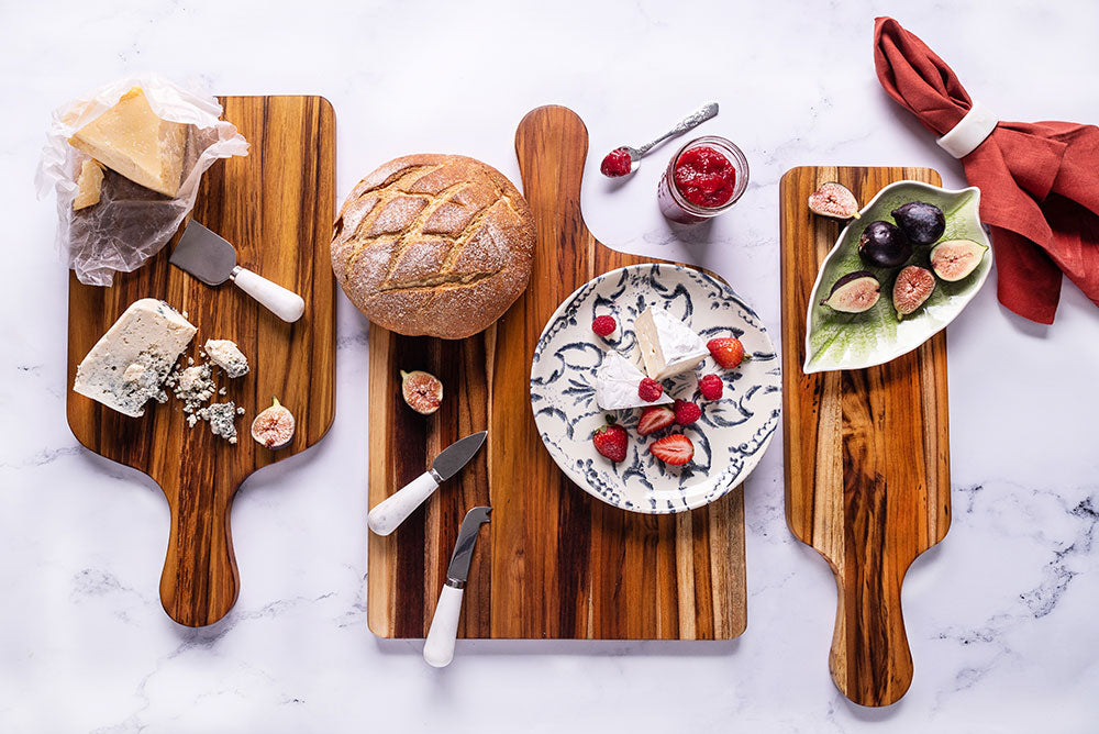 Teak wood marine paddle serving boards displaying cheese and fruit on a bright marble surface.