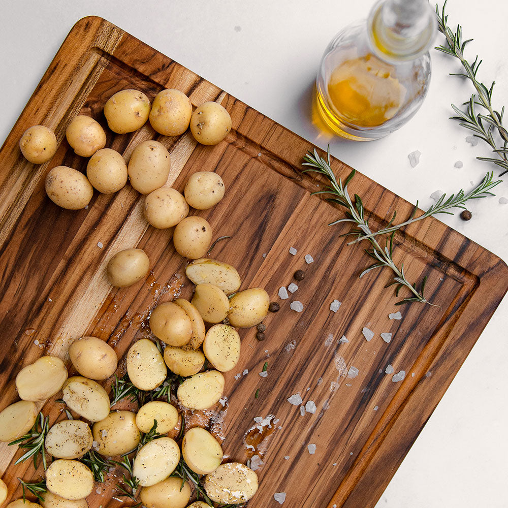 Thin edge grain teak cutting board with a juice canal, styled with baby potatoes, rosemary, and salt.