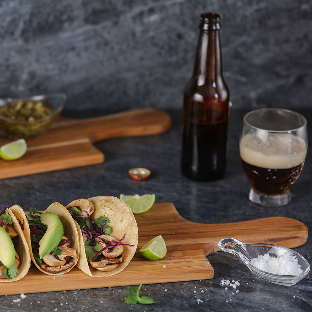 Teakwood table plank serving board with handle featuring mushroom tacos and limes on a dark surface.
