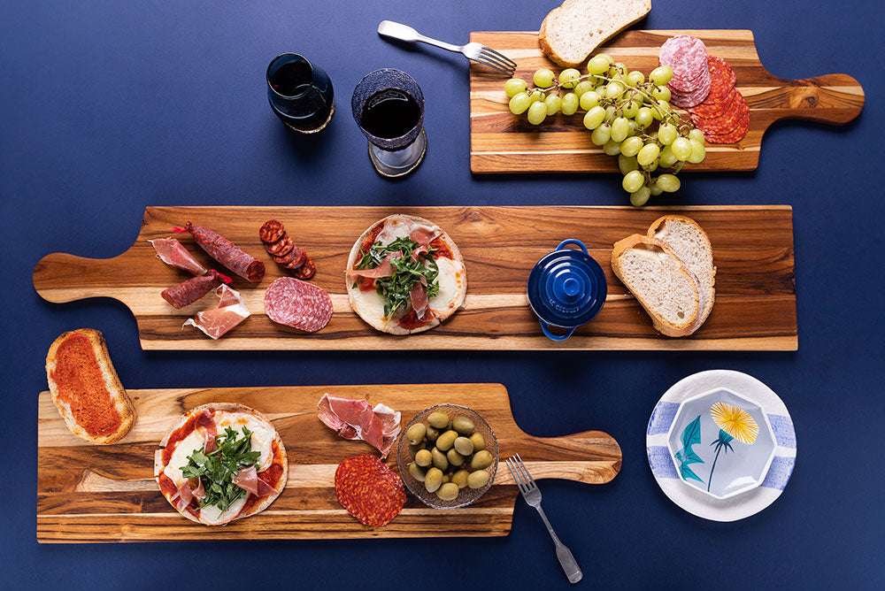 Teakwood table plank serving boards with handles featuring assorted charcuterie and grapes.