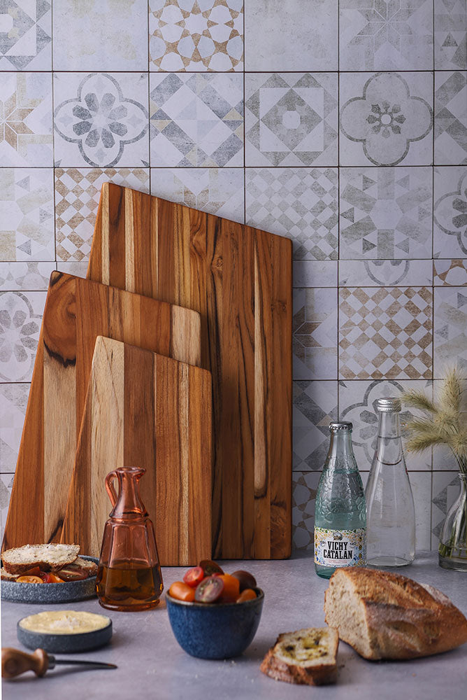 Trio of Geo Trapeze teakwood serving boards leaning against a patterned tile wall in a rustic kitchen scene.